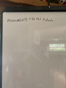 Whiteboard with one line handwritten in black: Assignments for PhD students